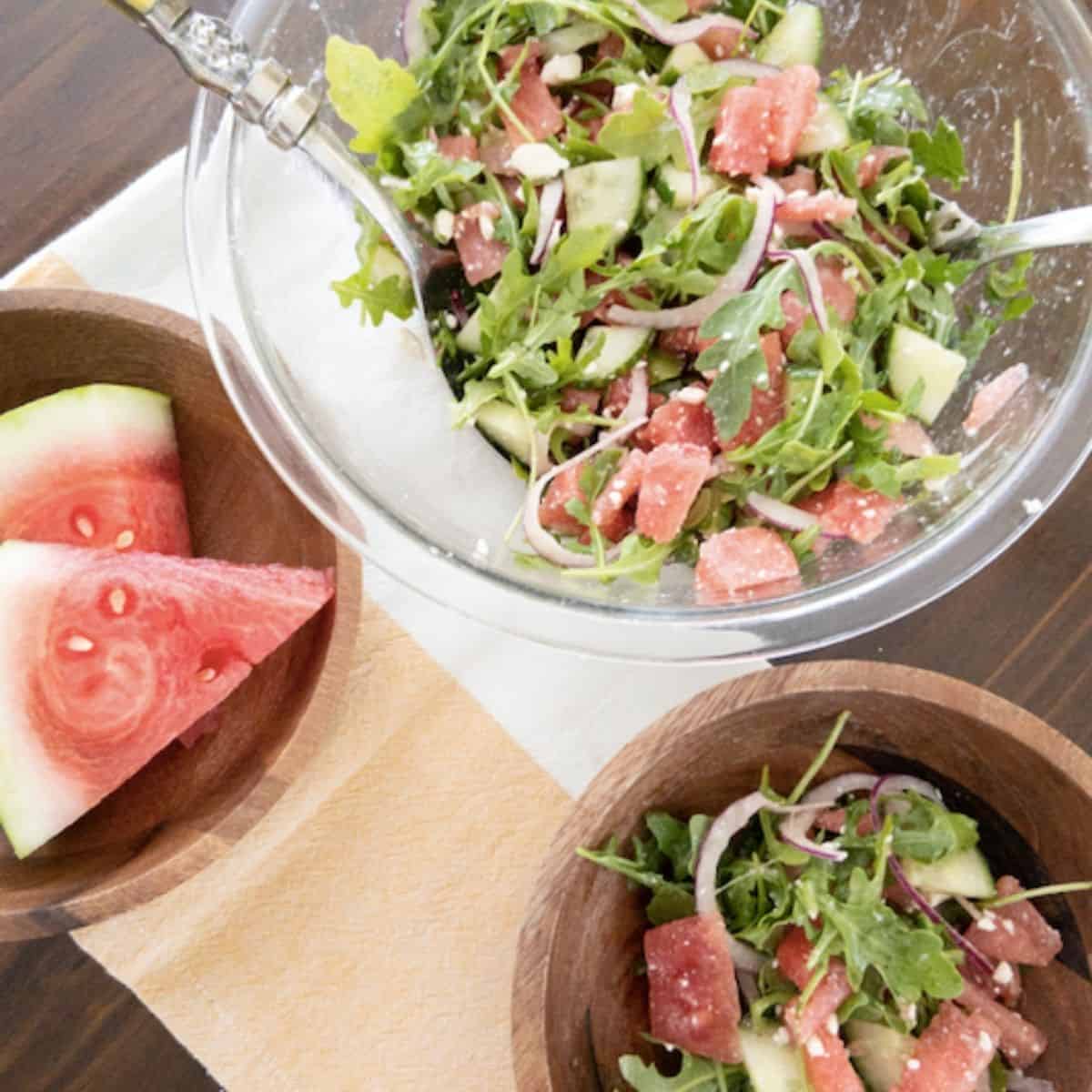 watermelon salad in glass bowl with serving tongs, a wooden bowl full of watermelon salad and watermelon slices on the side