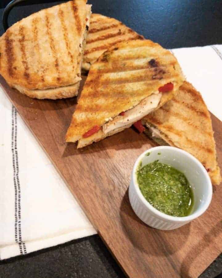 Pesto chicken sandwich cut in halves, on a cutting board with a side of basil pesto in a white bowl
