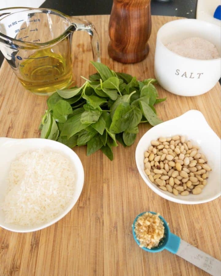All of the ingredients for basil pesto recipe including pine nuts, garlic, parmigiana reggiano cheese, basil, salt, olive oil