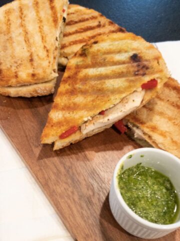 pesto chicken sandwiches cut into halves on wooden serving platter with a side of pesto
