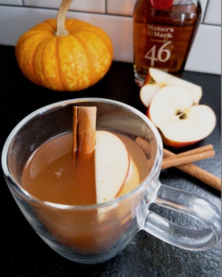 Apple cider bourbon cocktail in glass mug with makers mark in the background with apples and an orange pumpkin 