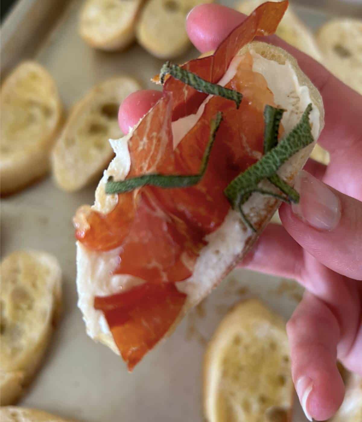 Hand holding crostini topped with goat cheese, prosciutto and sage.