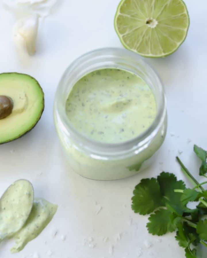 jar of avocado cream sauce surrounded by lime, cilantro, and an avocado