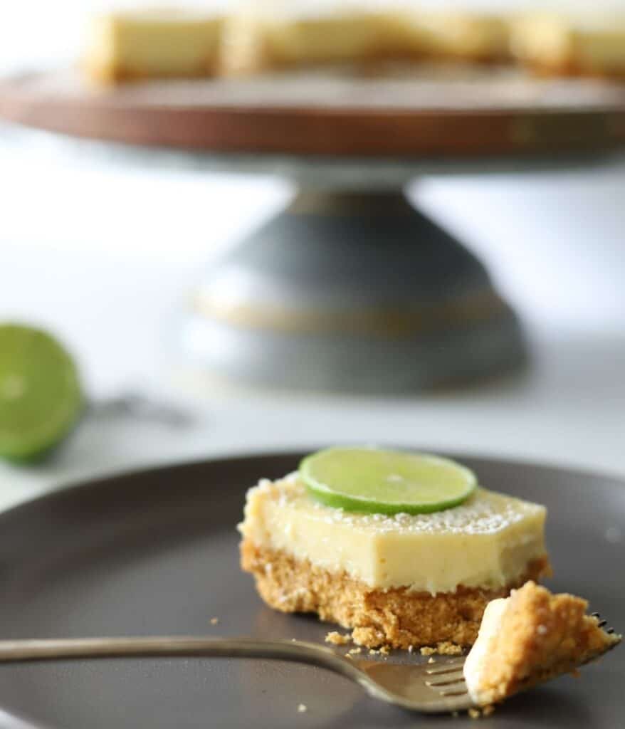 key lime pie bar on gray plate with a fork holding a bite of the dessert with cake plate in background holding more pie bars