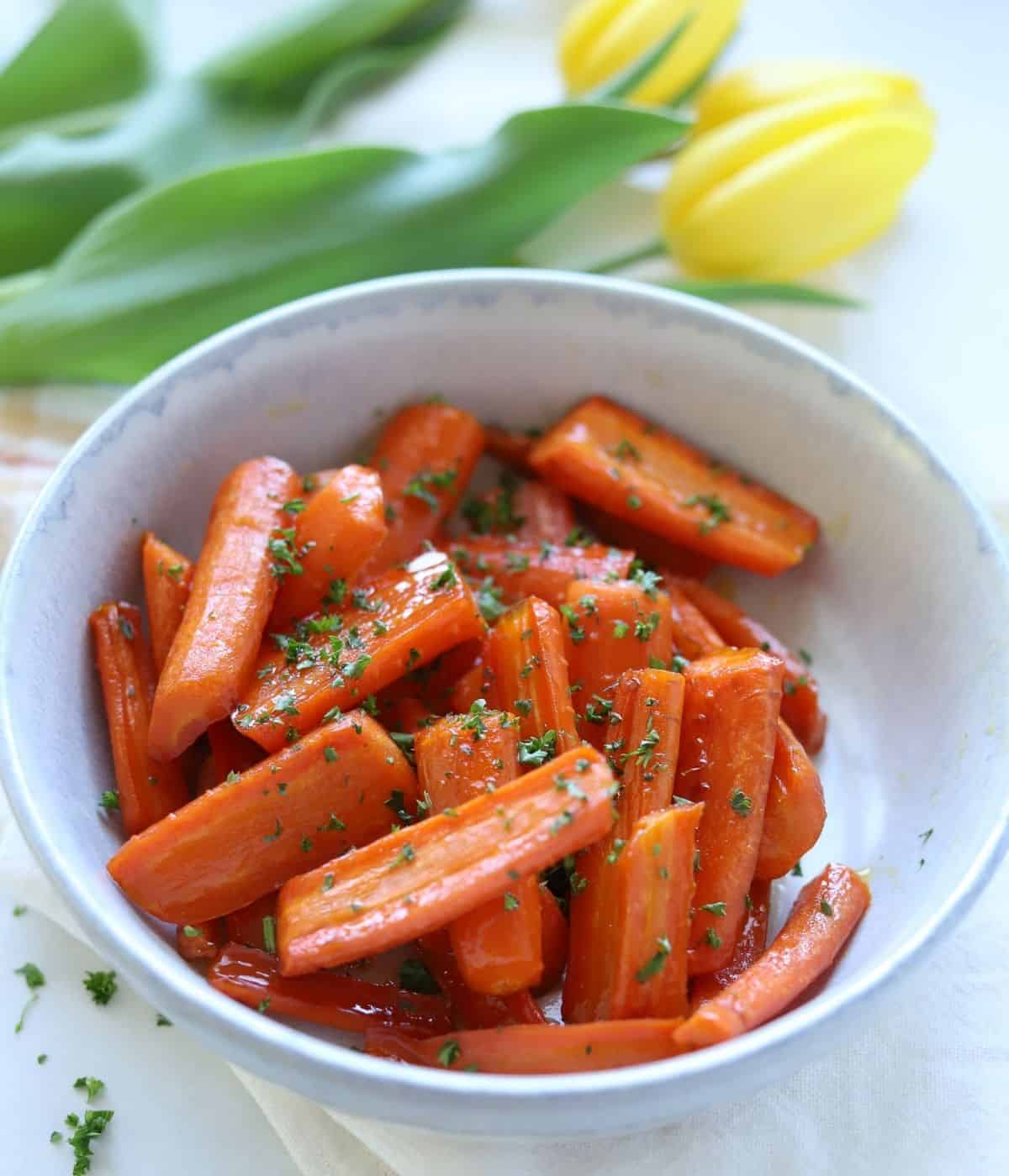 brown sugar honey glazed carrots in stoneware bowl topped with parsley with yellow tulips in background