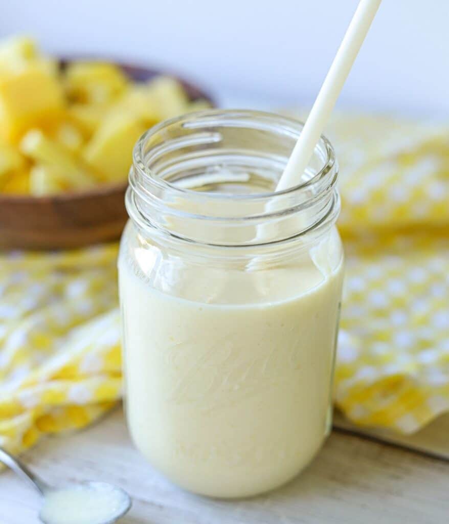 mason jar full of mango pineapple smoothie with yellow and white checkered napkin and wooden bowl full of pineapple and mango