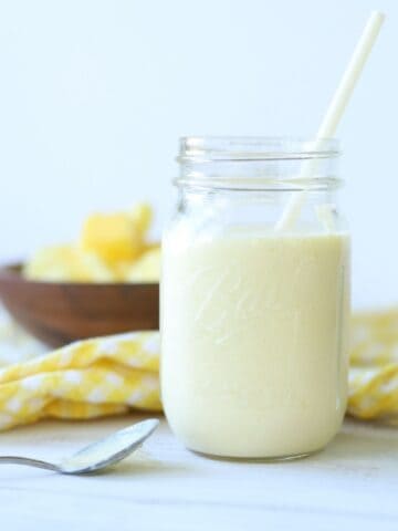 mango pineapple smoothie in a mason jar with a straw with yellow and white checkered napkin and wooden bowl full of mango and pineapple