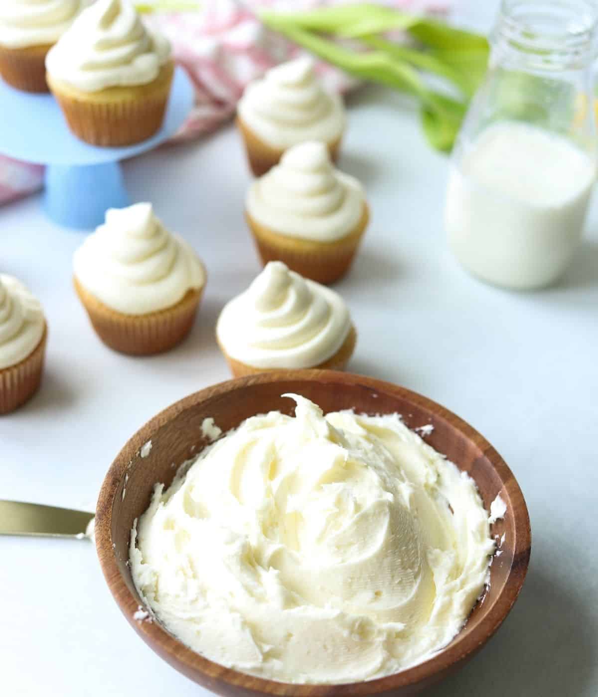 buttercream frosting in wooden bowl with cupcakes in background with milk and tulips