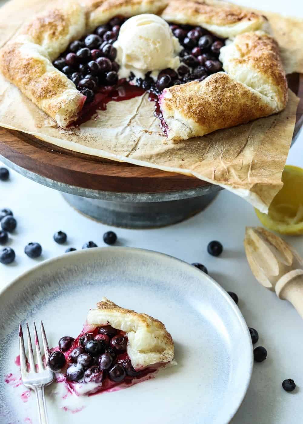 blueberry galette on a cake stand with scoop of vanilla ice cream on top and serving plate below it