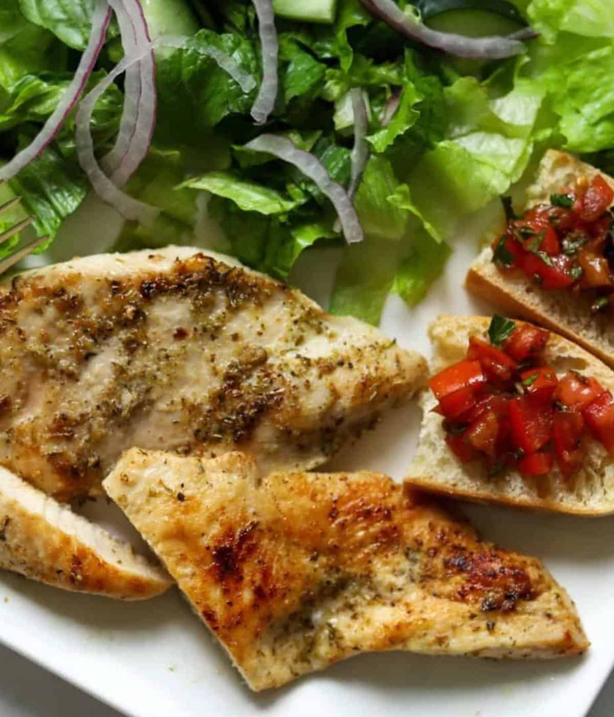 Sauteed chicken breast on plate with salad on side and bread with bruschetta.