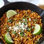 cast iron skillet filled with charred blackened corn topped with feta, cilantro and lime wedges
