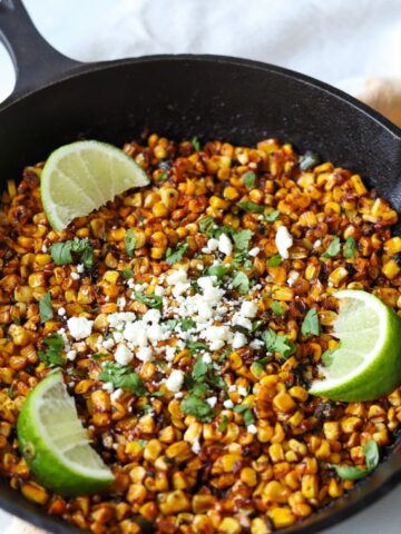cast iron skillet filled with charred blackened corn topped with feta, cilantro and lime wedges