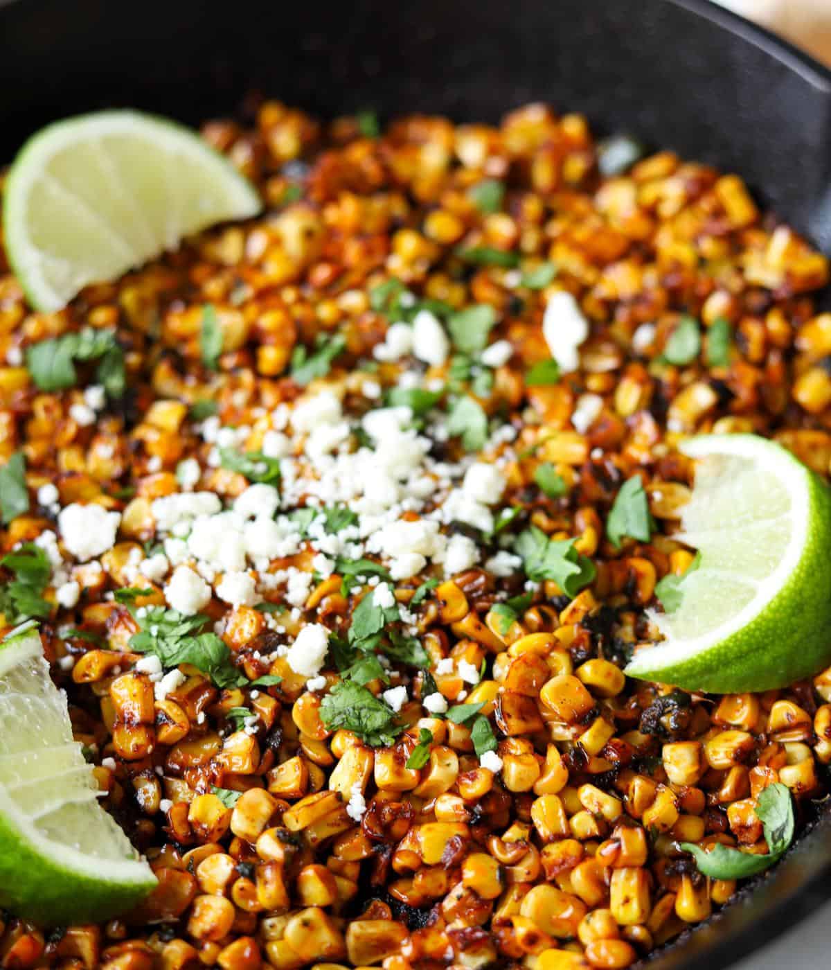 close up of blackened corn in skillet topped with cilantro, feta and lime wedges