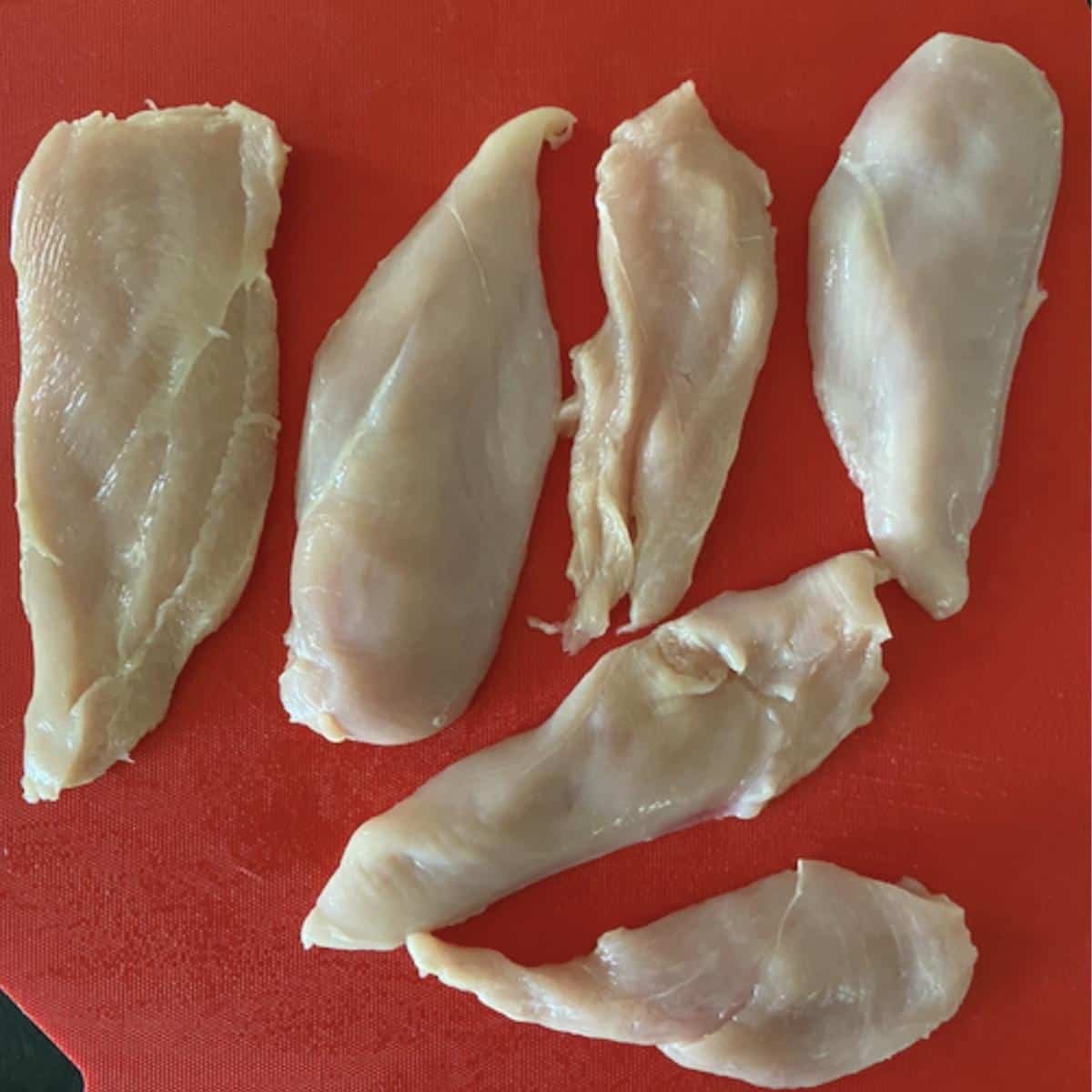 Chicken breast sliced in halves on red cutting board.