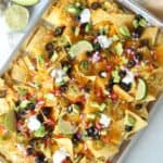 finished veggie nachos loaded with toppings on a cookie sheet with an avocado and lime on the side