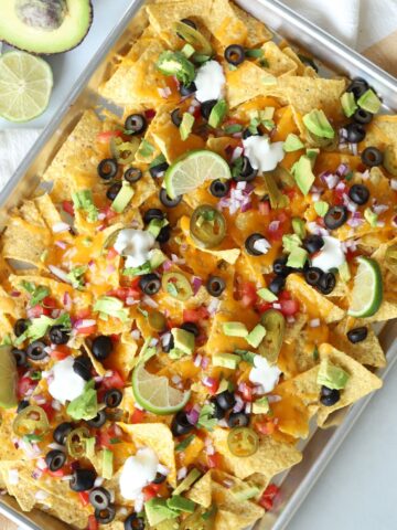finished veggie nachos loaded with toppings on a cookie sheet with an avocado and lime on the side