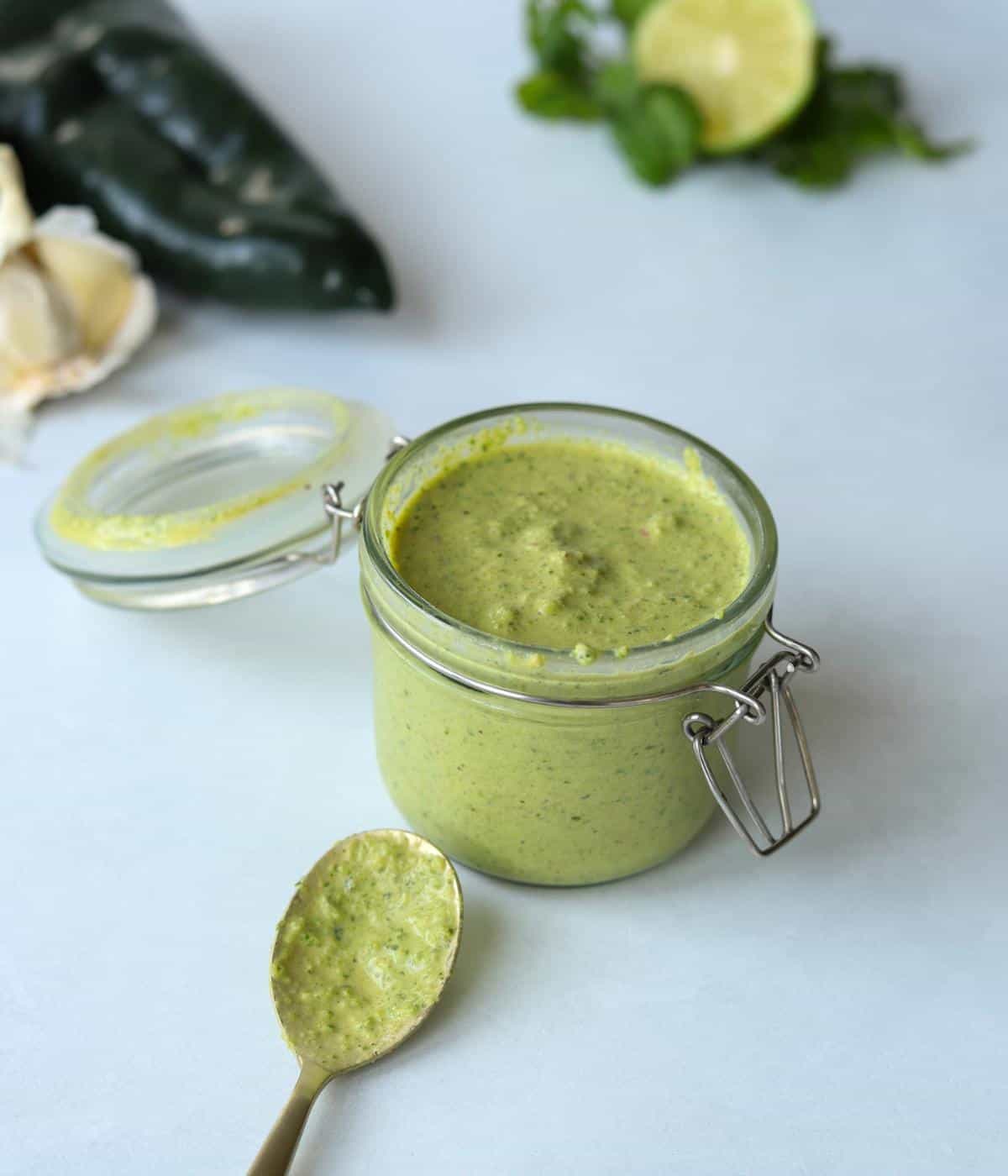 jar of poblano sauce with spoon laying in front and poblano and garlic in background