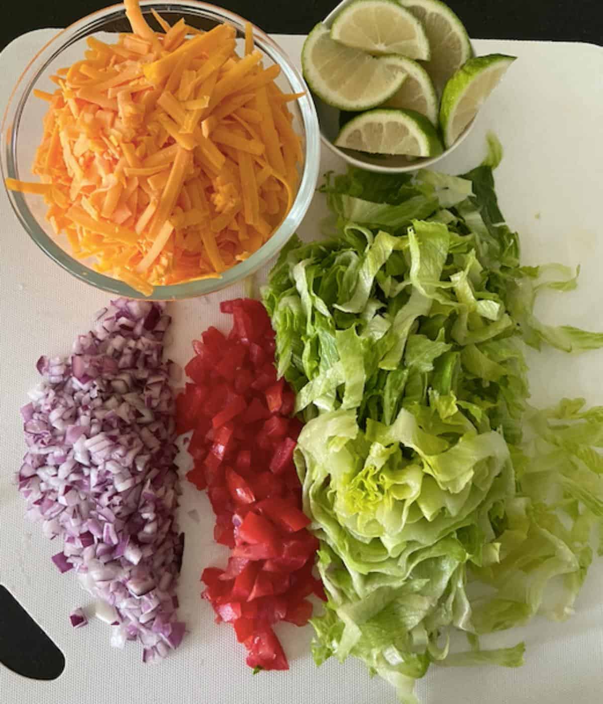 taco toppings on cutting board (lettuce, cheese, onion, tomato and limes)