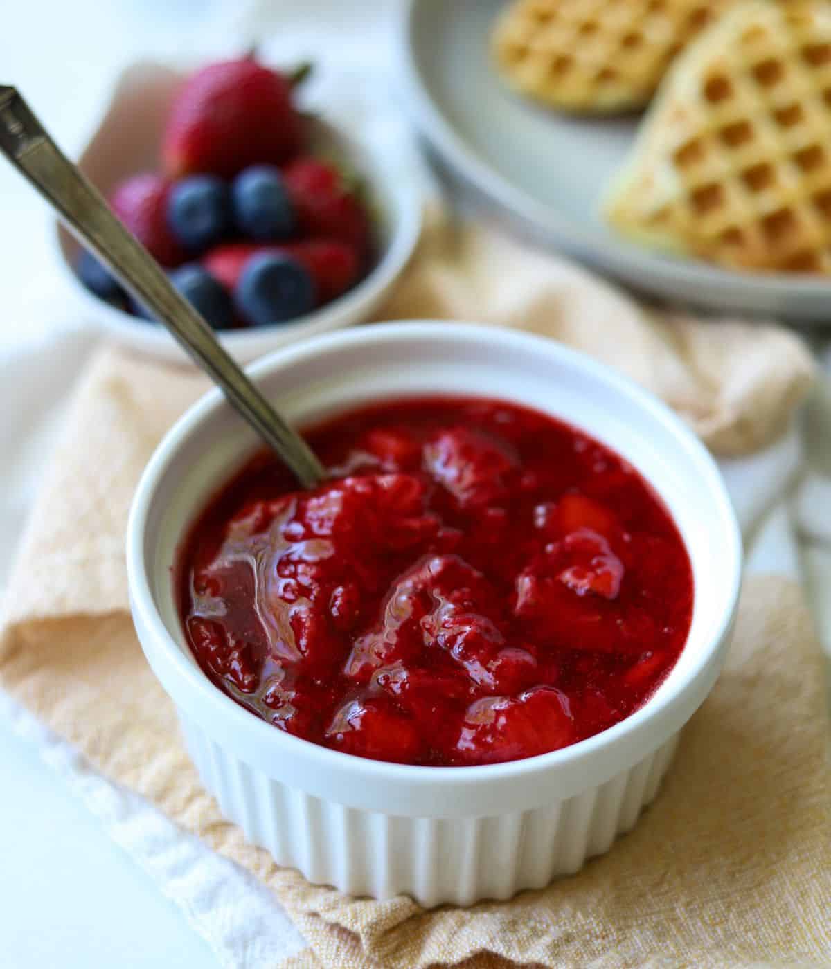 white bowl full of strawberry compote sauce with waffles and berries in background