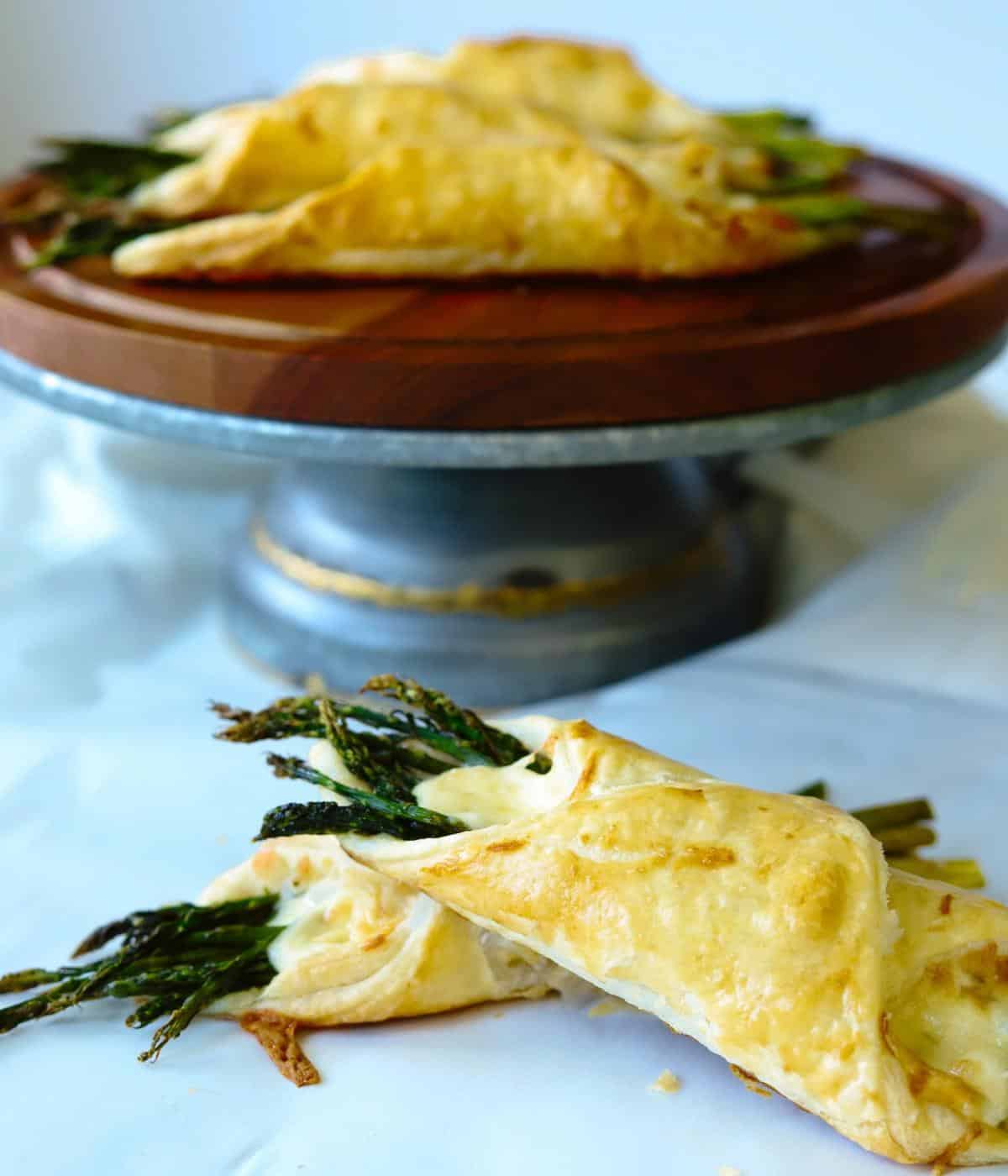 asparagus wrapped puff pastries on cake plate with two sitting in front.