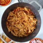 Dutch oven full of pulled chicken breast in bbq sauce with a side of slaw and beans.