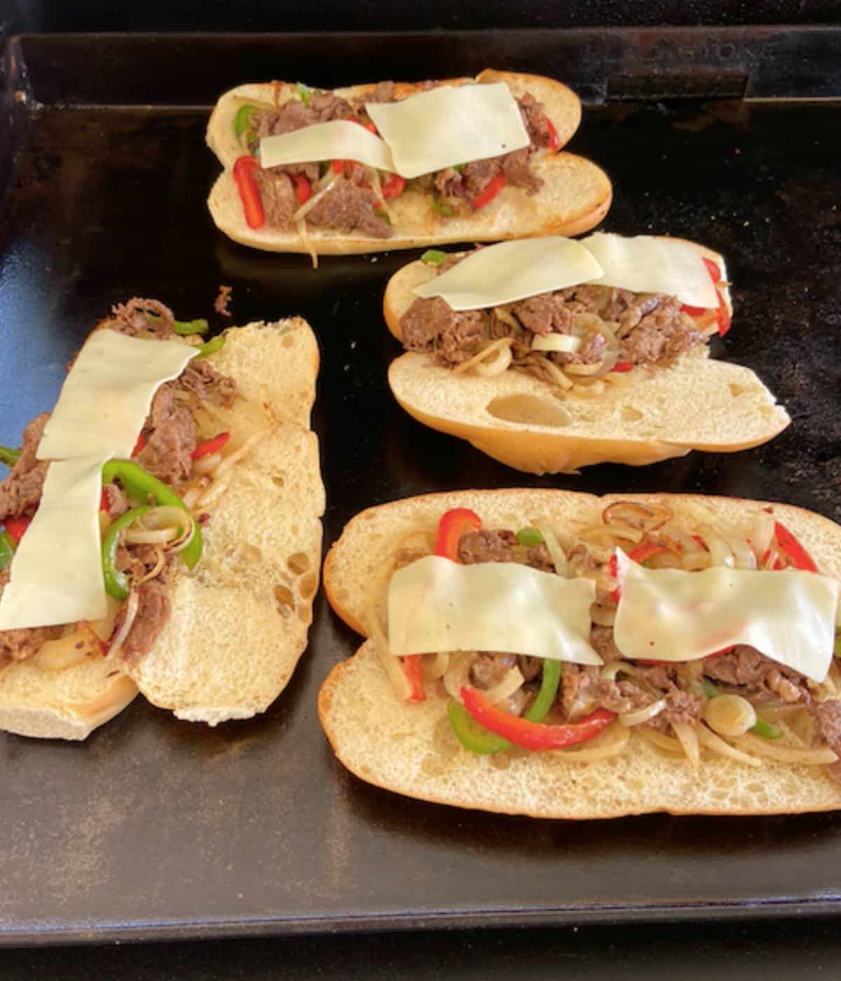 Philly Cheesesteaks topped with cheese on griddle.