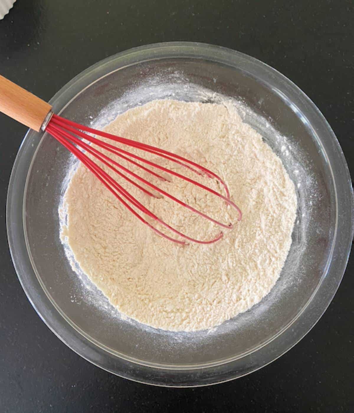 Dry ingredients in glass bowl with red whisk.
