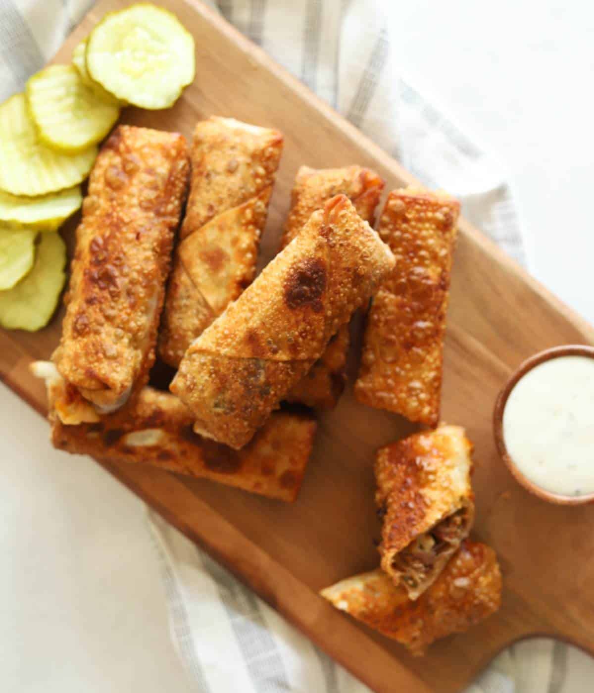 Egg rolls on wooden platter with ranch and pickles.