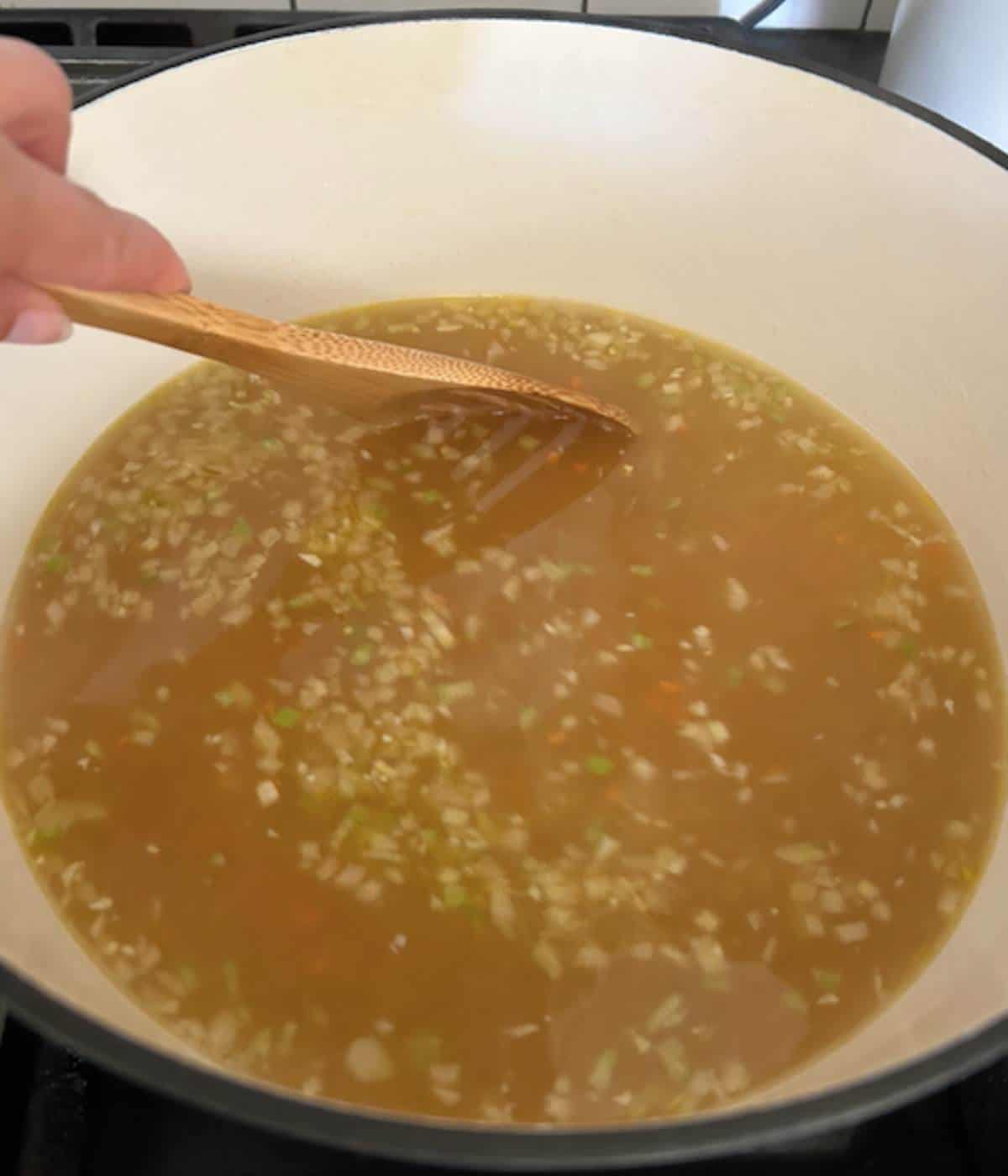 Hand stirring pastina into soup with wooden spoon.