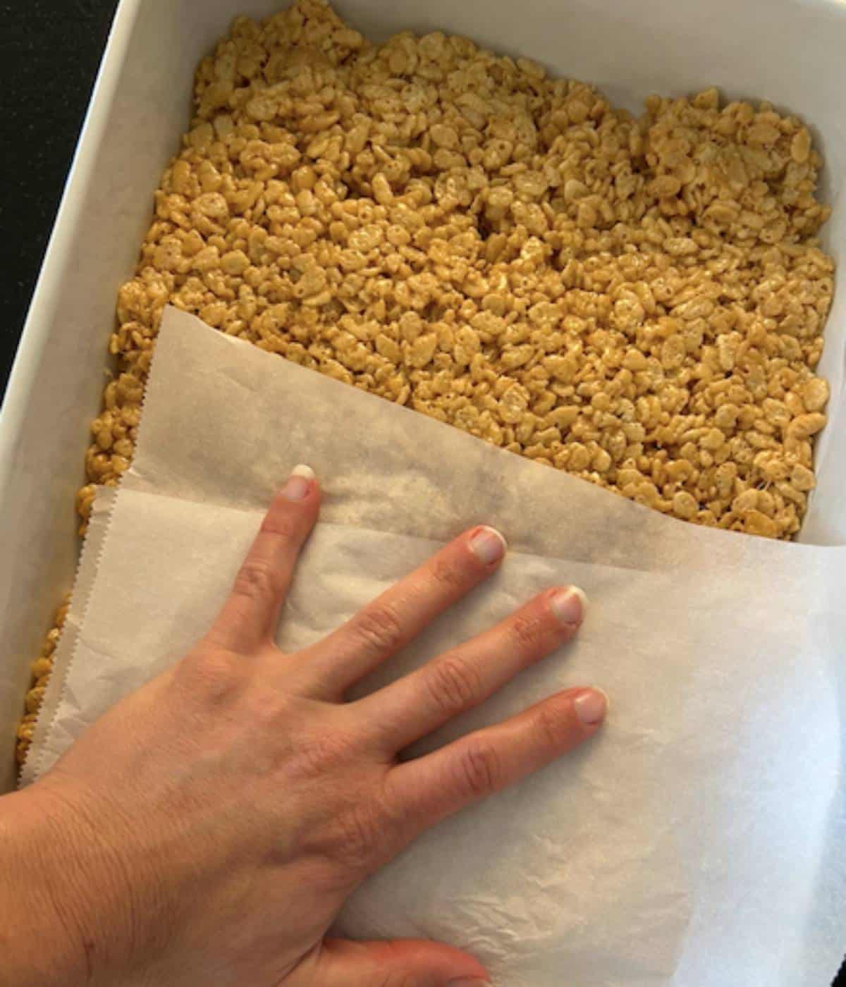 Hand with parchment paper flattening rice krispies in casserole dish.