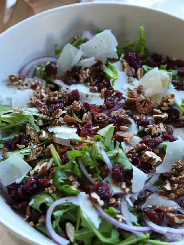 Spinach arugula salad topped with pecans, cranberries, parmesan cheese in large bowl.