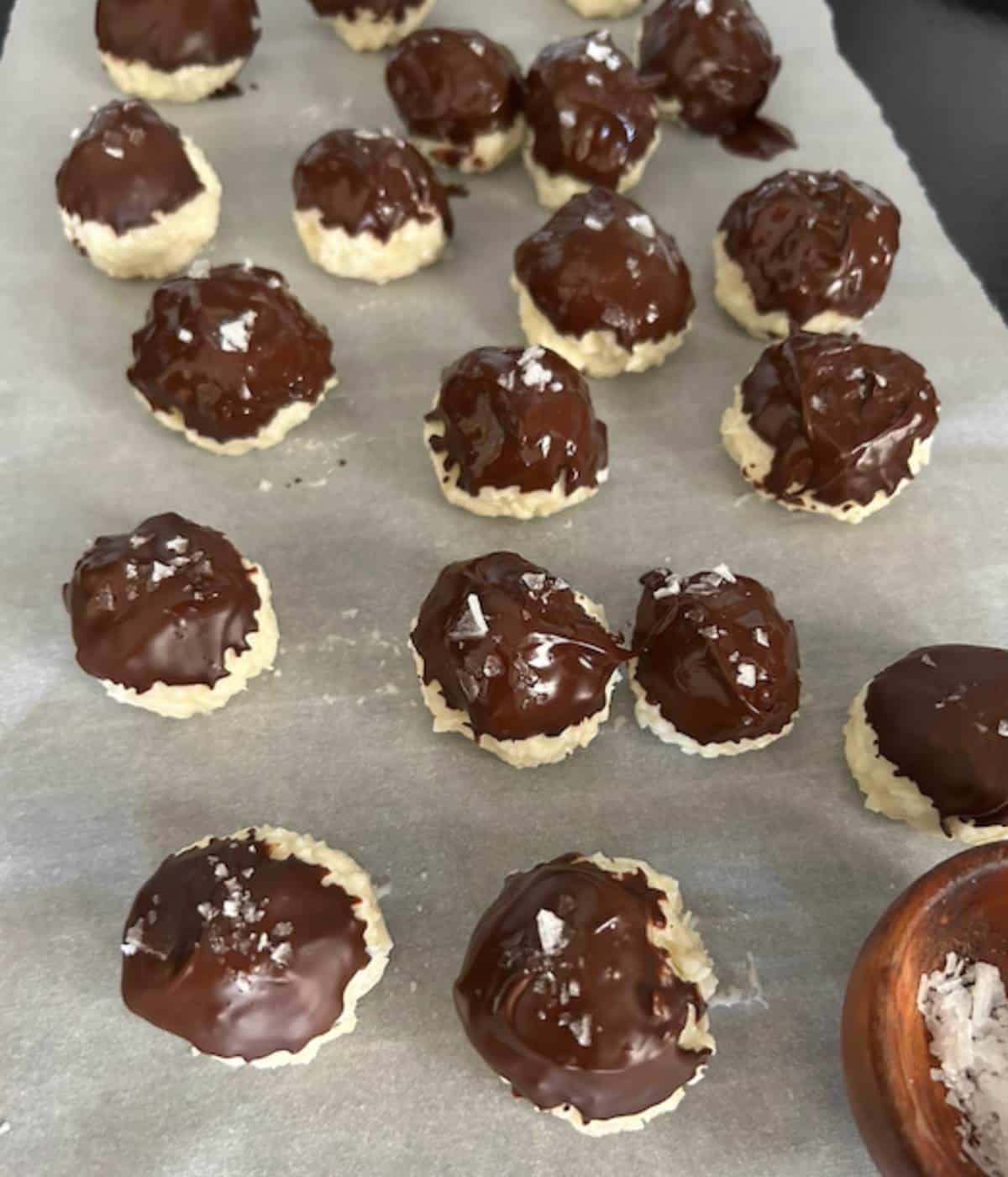 Coconut balls dipped in chocolate and topped with sea salt on parchment paper.