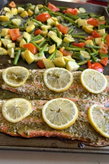 Pesto salmon with lemon slices and veggies on cookie sheet.